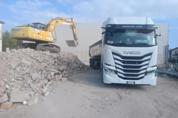 Camion con semirimorchio ribaltabile accanto a un escavatore e a un cumulo di materiali da demolizione in cantiere.
